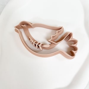 May include: A light brown dove-shaped cookie cutter with a sprig of leaves in its beak. The cookie cutter is made of plastic and is sitting on a white surface. The dove is a symbol of peace and is often used for baking.