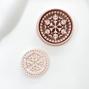 May include: Two round cookie stamps with snowflake designs. The larger stamp has a detailed snowflake and heart-shaped border. The smaller stamp has a simpler snowflake design with a dotted border. Both are light brown and sit on a white surface.