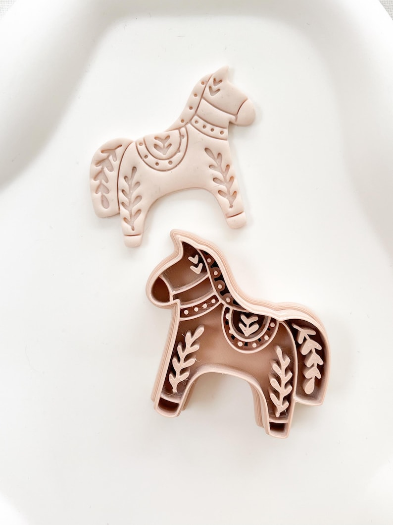 May include: Two horse-shaped cookie cutters. One is light pink, the other is brown. Both have decorative floral and dot patterns. The cutters are on a white surface.
