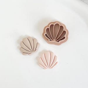 May include: Three seashell-shaped cookie cutters in varying shades of beige and pink. The largest cutter is a light brown color, and the other two are smaller, with one in a light beige and the other in a soft pink.