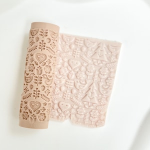 Könnte beinhalten: Eine beige Teigrolle mit einem geprägten Muster aus Vögeln, Herzen, Blumen und Bäumen. Die Rolle ist teilweise abgerollt und zeigt die gemusterte Oberfläche. Geeignet zum Erstellen dekorativer Muster auf Teig oder Ton.