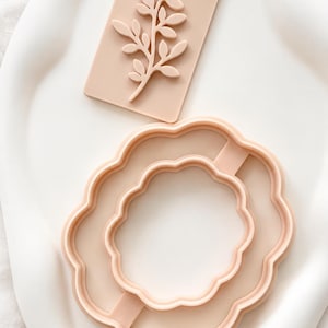 Op de afbeelding: Een licht perzikkleurige rechthoekige stempel met een verhoogd botanisch bladontwerp en een bijpassende licht perzikkleurige, geschulpte ronde koekjesvorm. Beide items zijn gemaakt van een glad, stevig materiaal met een matte afwerking.