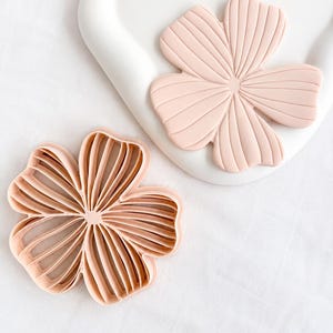 May include: A light peach-coloured flower-shaped cookie cutter with a hollow centre and raised interior ridges, paired with a matching light peach-coloured flower-shaped cutout piece that features embossed linear details on its surface.