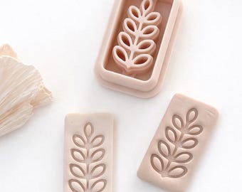 May include: A collection of light pink leaf-shaped cookie cutters and impressions. The cutters are rectangular with a detailed leaf design. The image shows the cutters and impressions on a white surface, ideal for baking and crafting.