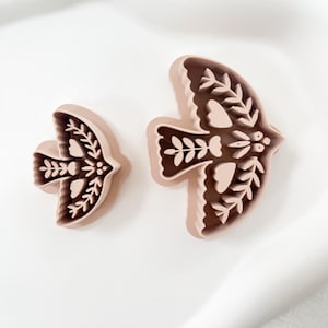 May include: Two bird-shaped cookie cutters. The cutters are light brown with a darker brown interior, featuring intricate floral and leaf designs. The cutters are placed on a white surface.