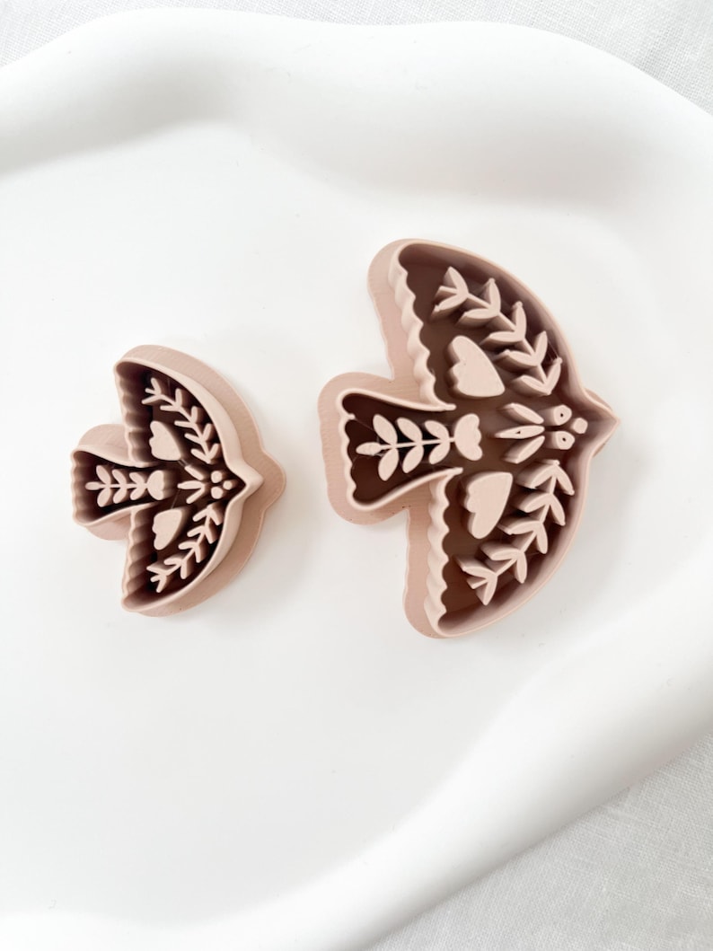 May include: Two bird-shaped cookie cutters. The cutters are light brown with a darker brown interior, featuring intricate floral and leaf designs. The cutters are placed on a white surface.