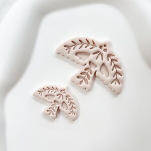 May include: Two light pink dove-shaped ornaments with leaf and heart cutouts. The larger dove is approximately 7.6 cm wide, and the smaller one is about 5 cm wide. Both are placed on a white, slightly curved surface.