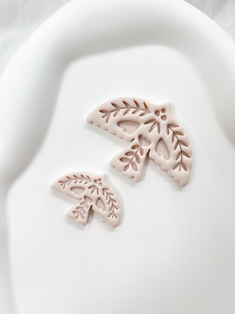 May include: Two light pink dove-shaped ornaments with leaf and heart cutouts. The larger dove is approximately 7.6 cm wide, and the smaller one is about 5 cm wide. Both are placed on a white, slightly curved surface.
