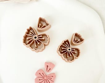 May include: A set of four pink plastic cookie cutters shaped like flowers. The cutters are arranged on a white surface. One of the cutters is filled with pink fondant.