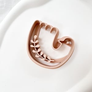 Puede incluir: Un cortador de galletas marrón claro en forma de mano con un diseño floral. El cortador es de plástico y está diseñado para hornear. La forma de la mano tiene un diseño curvo con una pequeña rama floral.
