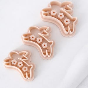 May include: Three light pink bunny-shaped cookie cutters with flower details. The cutters are made of plastic and are arranged on a white surface. The bunny cutters are for baking and crafting.