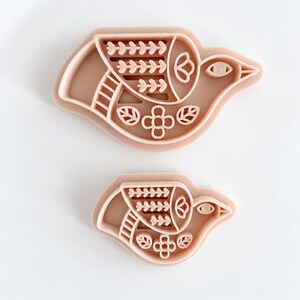 Puede incluir: Dos cortadores de galletas en forma de pájaro de color melocotón claro con intrincados detalles en relieve. El diseño presenta un pájaro estilizado con un patrón de hojas en el ala, un motivo floral central y un ojo pequeño, en plástico mate.