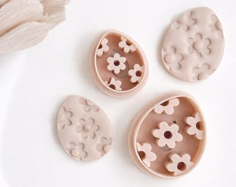 May include: Four light pink egg-shaped cookie cutters and embossed cookies with a floral design. The cutters have a raised edge and the cookies have a detailed flower pattern. The items are arranged on a white surface.