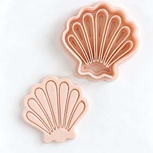 Pode incluir: Um conjunto de cortadores de biscoitos de plástico rosa claro em forma de concha. O conjunto inclui uma peça de carimbo sólida com cristas em relevo e uma moldura de corte externa separada com detalhes internos para criar um design de concha.