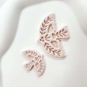 May include: Two light pink dove-shaped ornaments with intricate cut-out leaf and heart designs. The ornaments are in different sizes, with the larger one positioned above the smaller one. The background is a white, slightly curved surface.