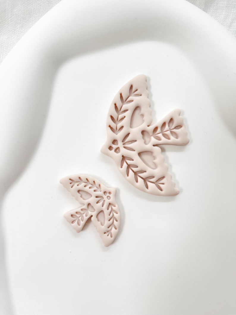 May include: Two light pink dove-shaped ornaments with intricate cut-out leaf and heart designs. The ornaments are in different sizes, with the larger one positioned above the smaller one. The background is a white, slightly curved surface.