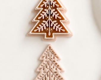 May include: Two light peach-colored plastic cookie cutters shaped like Christmas trees. The top cutter is a two-piece set with a brown interior, while the bottom cutter is a single piece featuring a detailed, intricate cutout pattern of branches.
