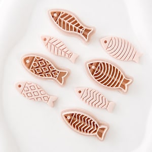Puede incluir: Un conjunto de siete cortadores de galletas en forma de pez de color rosa claro. Cada cortador presenta un patrón interno distinto, incluyendo rejillas geométricas, rayas verticales y diseños de espinas, con un borde elevado.