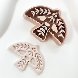 May include: Two bird-shaped cookie cutters. One is a solid, light brown colour with intricate leaf and heart cutouts. The other is a lighter shade of brown with a detailed leaf and heart design. Both are on a white surface.