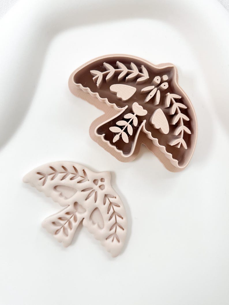 May include: Two bird-shaped cookie cutters. One is a solid, light brown colour with intricate leaf and heart cutouts. The other is a lighter shade of brown with a detailed leaf and heart design. Both are on a white surface.
