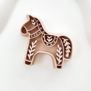 May include: A light brown horse-shaped cookie cutter with decorative floral and dot patterns. The cookie cutter is made of plastic and is designed with a detailed, folk art style. The horse has a rounded body and a curved neck.