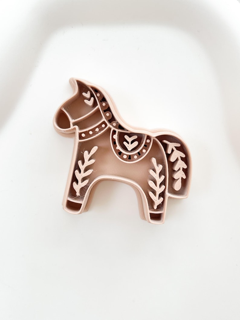 May include: A light brown horse-shaped cookie cutter with decorative floral and dot patterns. The cookie cutter is made of plastic and is designed with a detailed, folk art style. The horse has a rounded body and a curved neck.