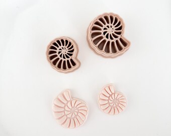 May include: Four seashell-shaped cookie cutters in varying sizes. Two are a darker tan color, and two are a lighter pink. The cutters are arranged on a white surface, showcasing their spiral design.