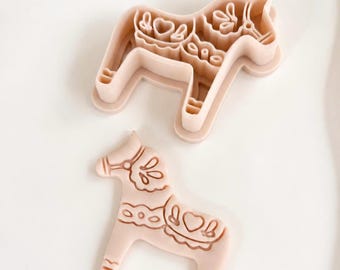 May include: A light pink horse-shaped cookie cutter and a matching stamp featuring intricate embossed details including a heart, floral patterns, and a bridle. The set consists of two separate pieces designed to create a detailed horse-shaped cookie.