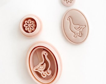 May include: A collection of pink cookie cutters in various shapes. The cutters include a flower and a goose design. The goose design is in an oval shape. The cookie cutters are made of plastic and are on a white background.