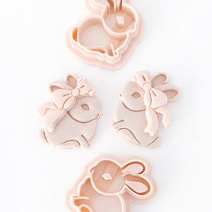 Pode incluir: Quatro cortadores de biscoitos em forma de coelho rosa claro. Dois cortadores têm um laço na cabeça do coelho. Os cortadores são feitos de plástico e são projetados para assar. Os coelhos estão em poses diferentes.