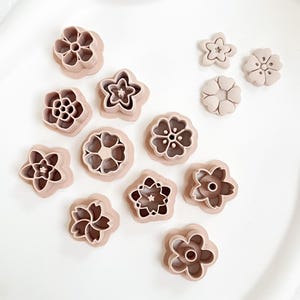 Puede incluir: Una colección de cortadores de galletas con forma de flor de color marrón claro en varios diseños. Los cortadores están dispuestos sobre una superficie blanca, mostrando diferentes patrones florales. Estas herramientas son ideales para hornear y manualidades.
