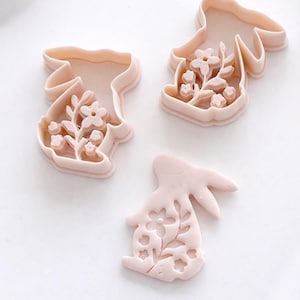 Pode incluir: Três cortadores de biscoitos em forma de coelho rosa claro com desenhos florais. Os cortadores são feitos de plástico e estão dispostos sobre uma superfície branca. Os detalhes florais são visíveis dentro das formas de coelho, sugerindo um tema primaveril ou de Páscoa.
