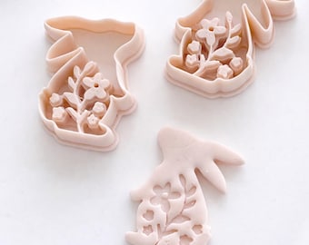 May include: Three light pink bunny-shaped cookie cutters with floral designs. The cutters are made of plastic and are arranged on a white surface. The floral details are visible within the bunny shapes, suggesting a spring or Easter theme.
