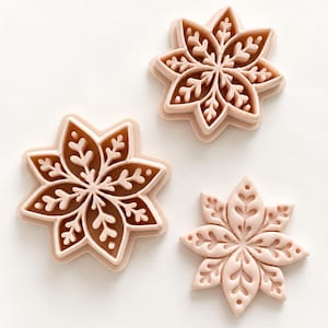 May include: Three light pink plastic cookie cutters shaped like intricate snowflakes. Two of the cutters have a brown interior base, while the third cutter is solid light pink, all featuring detailed embossed patterns of branching petals.