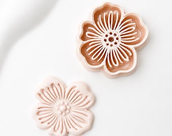 May include: A light peach-colored flower-shaped cookie cutter and a matching embossed flower shape. The cutter features a detailed petal and stamen design with a raised outer rim, while the embossed piece shows the corresponding floral pattern.