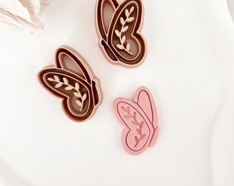 May include: Three butterfly-shaped cookie cutters. Two are brown, and one is pink. Each cutter has a leaf design inside the wings. The cutters are on a white surface.