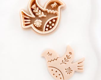 May include: A light peach-colored bird-shaped cookie cutter and a matching stamped bird-shaped cookie piece. The cutter features intricate cutouts of a floral branch, a star-like flower, and a series of small circles on the bird's wing and body.