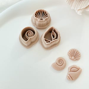 May include: A collection of tan seashell-shaped cookie cutters in various designs, including a scallop, spiral, and conch shell. The cutters are arranged on a white surface, with a soft, neutral background.