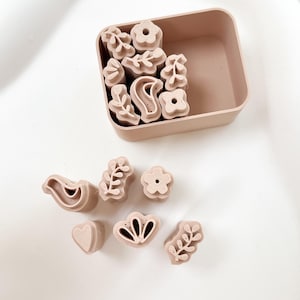 May include: A set of light brown floral and nature-themed stamps, including flowers, leaves, and a bird, are displayed in a matching square container. Several stamps are outside the container. The stamps are made of a solid material.