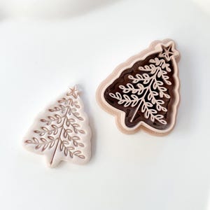 Könnte beinhalten: Zwei Ausstecher in Form eines Weihnachtsbaums. Einer ist hellbeige mit einem detaillierten Baumdesign. Der andere ist ein mehrschichtiges Design mit einem dunkelbraunen Innenraum und einem beigefarbenen Äußeren, ebenfalls mit einem Baumdesign.