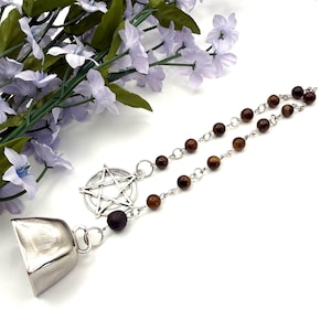 Puede incluir: Un dije de campana plateada con un colgante de pentagrama y una cadena de cuentas con cuentas marrones. El dije se muestra sobre un fondo blanco con flores moradas. La campana tiene una superficie pulida y reflectante.