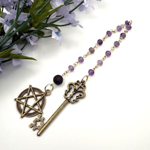 May include: A bronze-toned pendant featuring a pentagram and a key, connected by a strand of purple beads. The key has an ornate design. The pendant is likely a piece of jewelry or a decorative item.