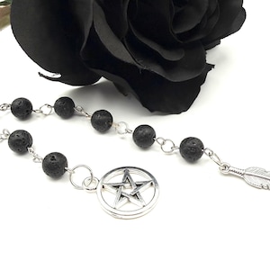 Op de afbeelding: Een zwarte lavastenen rozenkrans met zilverkleurige accenten. De rozenkrans heeft een pentagram bedel en een veervormige hanger. Een zwarte roos staat op de achtergrond. De rozenkrans ligt op een witte ondergrond.