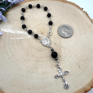 Könnte beinhalten: Ein silberfarbenes Rosenkranzarmband mit schwarzen Lavaperlen und einem dekorativen Kreuzanhänger. Das Armband hat ein kleines Medaillon und einen Verschluss. Eine Münze liegt neben dem Armband als Größenreferenz.