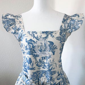 May include: A white and blue toile dress displayed on a mannequin. The dress features a square neckline, short sleeves, and a bow at the waist. The fabric is patterned with pastoral scenes in blue, with a central medallion.