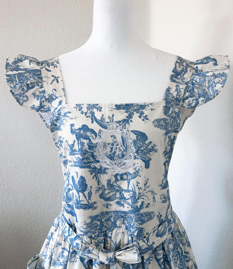 May include: A white and blue toile dress displayed on a mannequin. The dress features a square neckline, short sleeves, and a bow at the waist. The fabric is patterned with pastoral scenes in blue, with a central medallion.