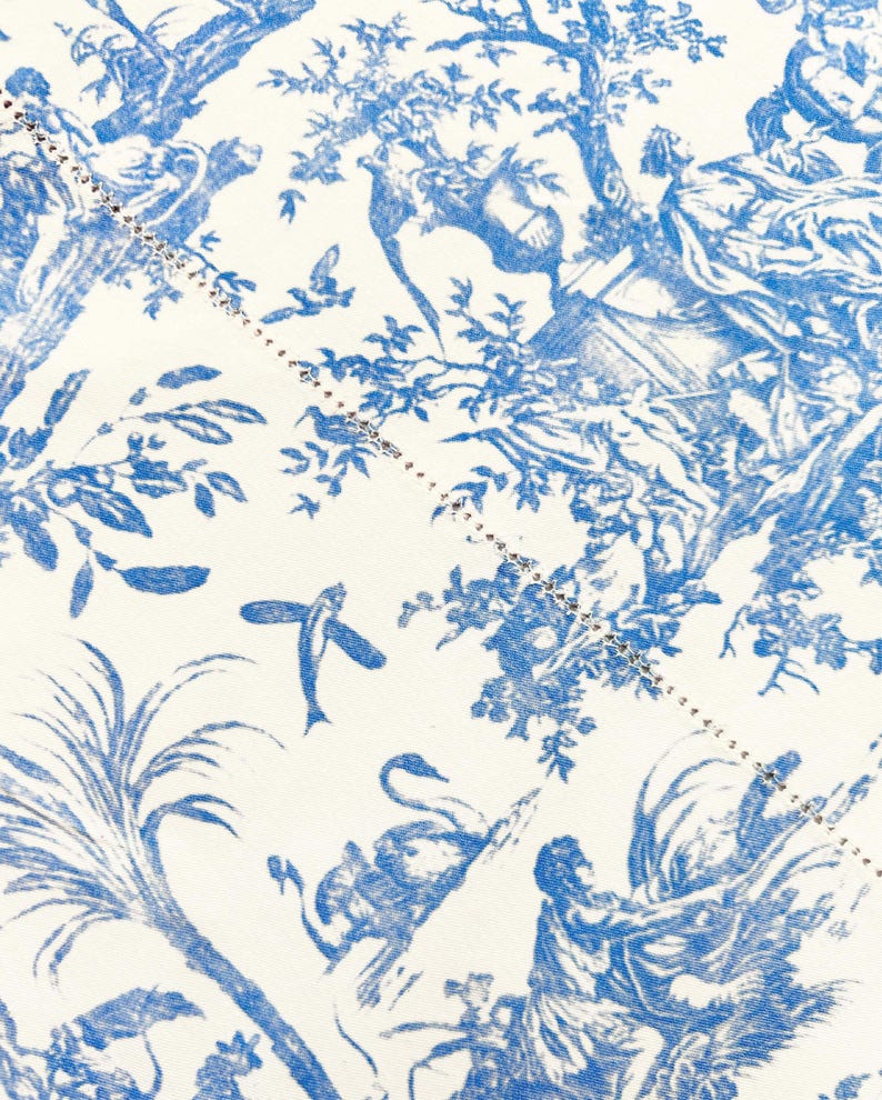May include: Close-up of a fabric with a blue and white toile pattern. The design features classical figures, trees, birds, and other natural elements. A dotted line with small embellishments runs diagonally across the fabric.
