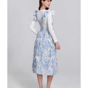 May include: A white long-sleeved top with a blue and white floral print dress. The dress has suspender straps and a midi-length skirt. The outfit is completed with white sandals. The overall style is feminine and elegant.