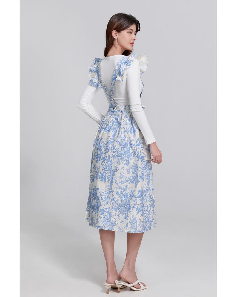 May include: A white long-sleeved top with a blue and white floral print dress. The dress has suspender straps and a midi-length skirt. The outfit is completed with white sandals. The overall style is feminine and elegant.