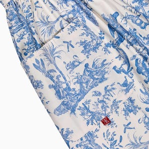 May include: White and blue toile fabric with a pocket. The fabric features a detailed pastoral scene with figures, trees, and birds. A red tag with a floral design is visible on the pocket. The overall design is reminiscent of classic French country style.
